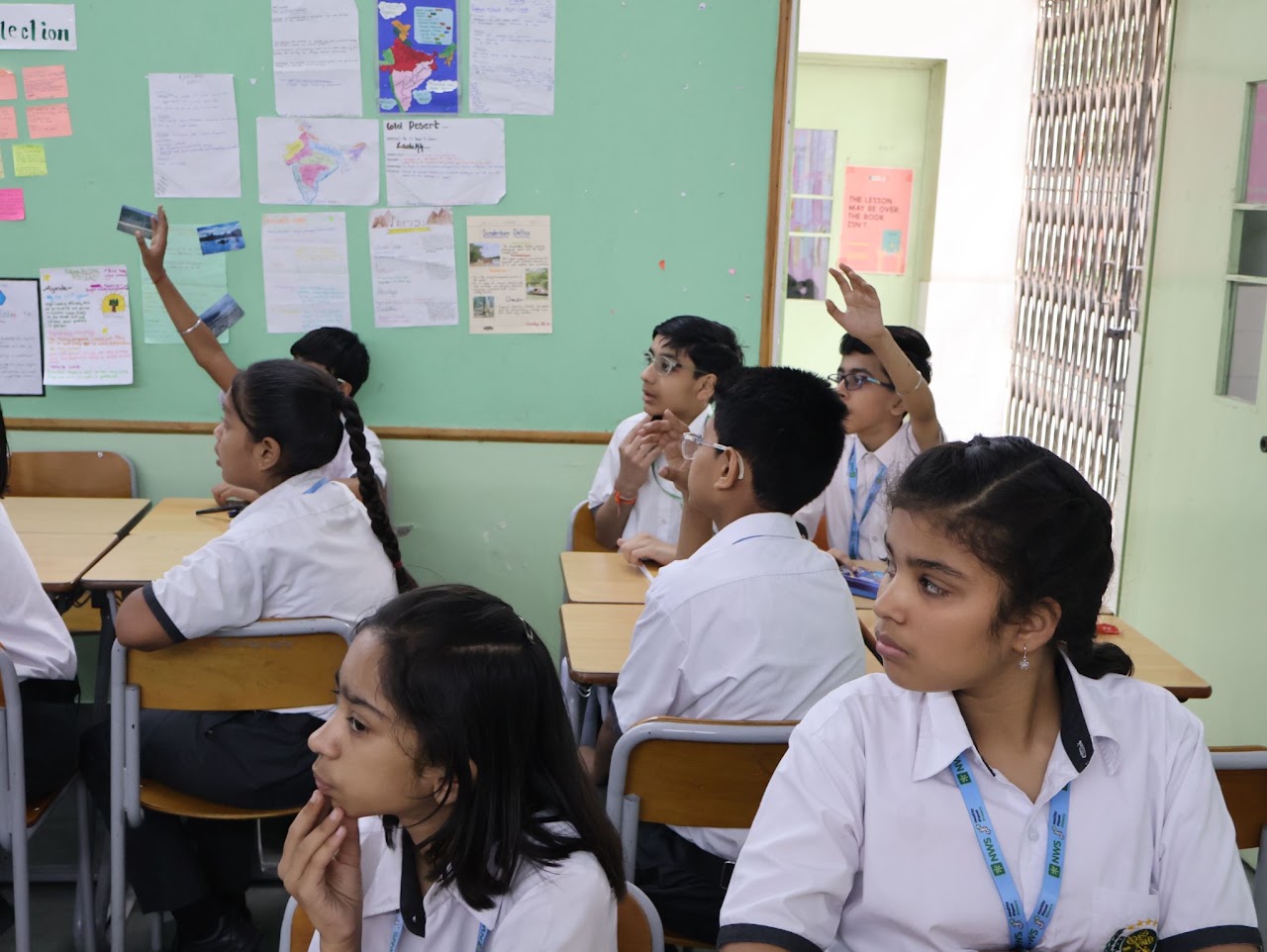 Students at their desks in a classroom, several with hands raised